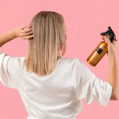 Une femme en peignoir blanc tient un flacon doré, prêt à utiliser du peroxyde d'hydrogène pour éclaircir ses cheveux blonds.