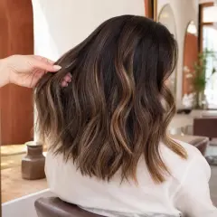 Une main ajuste une chevelure brune aux reflets subtils de balayage caramel, dans un salon de coiffure lumineux.