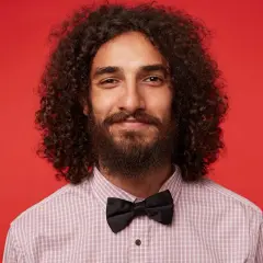 Homme souriant avec de longs cheveux bouclés, barbe et nœud papillon noir, sur fond rouge.