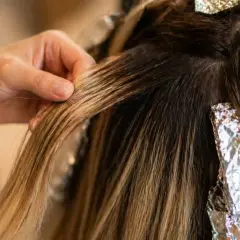 Main d'une coiffeuse travaillant sur des mèches de cheveux blonds, avec des feuilles d'aluminium. Le **foilyage vs balayage** est en cours.