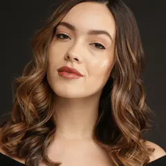 Une femme aux cheveux bruns ondulés, avec des reflets blonds, pose pour une photo. Elle a un maquillage naturel et un sourire subtil. Elle semble avoir réussi à faire un balayage maison.