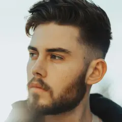 Jeune homme avec une barbe courte et une coupe de cheveux dégradée, style taper fade. Il regarde au loin, le visage éclairé par une lumière douce.