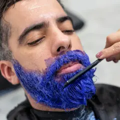 Un homme se fait teindre la barbe en bleu vif par une main tenant une brosse.