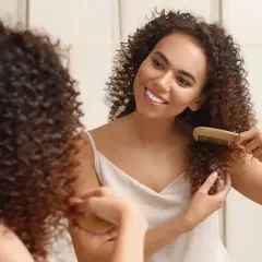 Une jeune femme souriante se regarde dans un miroir, peignant ses cheveux bouclés courts avec un peigne en bois.