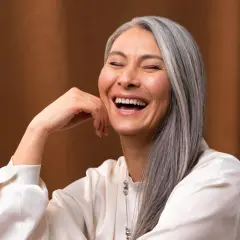 Une femme rit, sa main posée sur sa joue. Ses cheveux gris argenté ont des mèches de couleur subtiles, ajoutant une touche de modernité à son look.