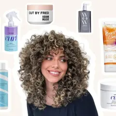 Une femme aux cheveux bouclés souriante, entourée de produits pour une routine cheveux bouclés : sprays, masques, gels et conditionneurs.