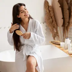 Une femme souriante en peignoir blanc se brosse les cheveux longs et bruns, un geste doux pour nourrir ses cheveux.