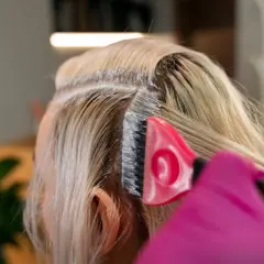 Application d'un produit super éclaircissant ou décoloration sur les racines des cheveux blonds. Une brosse rose et des gants roses sont visibles.