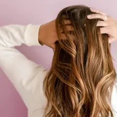 Une femme ajuste sa chevelure brune ondulée, révélant des reflets dorés sous la lumière.