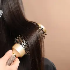Une main utilise une brosse ronde pour coiffer des cheveux bruns, tandis qu'un sèche-cheveux souffle de l'air. Le brushing boucle est en cours.