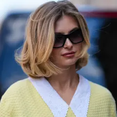 Une femme aux cheveux mi-longs blonds avec des reflets miel, portant des lunettes de soleil oversize et un pull jaune pâle à col blanc brodé. Une belle idée couleur cheveux mi long.