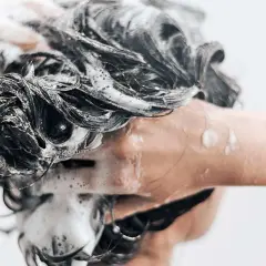 Une main savonne des cheveux sombres et mouillés, créant une mousse abondante. Une question se pose : peut-on se laver les cheveux au savon de Marseille ?