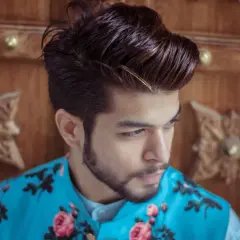 Un jeune homme aux cheveux longs et coiffés, portant une veste bleue à fleurs roses, se tient devant une porte en bois sculpté.