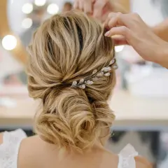 Une mariée se fait coiffer un chignon bas flou, orné d'une barrette scintillante. Les mains d'une coiffeuse ajustent les mèches.