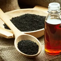 Une cuillère en bois remplie de graines de nigelle noire, à côté d'un bol de graines et d'une bouteille d'huile ambrée, parfaite pour l'huile de nigelle cheveux.