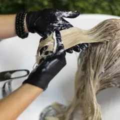 Application d'une patine pour foncer les cheveux blonds. Les mains gantées de noir appliquent le produit mèche par mèche.