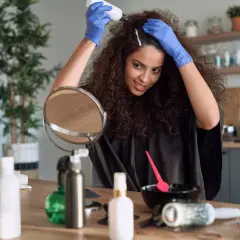 Une femme souriante fait sa coloration cheveux maison, appliquant le produit avec des gants bleus. Un miroir et des accessoires de coiffure sont sur la table.