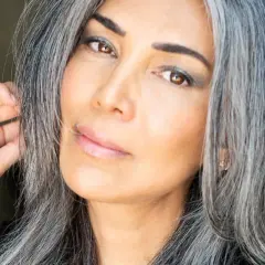 Une femme avec des cheveux gris mi-courts, le regard intense, touche doucement sa chevelure.