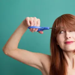 Une femme aux cheveux roux tente de comment couper une frange avec des ciseaux bleus.