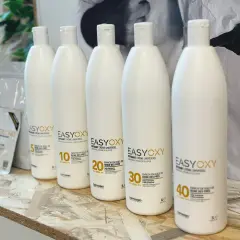 Cinq bouteilles d'oxydant seul pour éclaircir, de différentes forces (5, 10, 20, 30, 40 volumes), sur une étagère en bois.