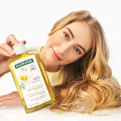Une femme aux cheveux blonds tient un flacon de shampoing Klorane à la camomille, un produit pour cheveux éclaircissant.