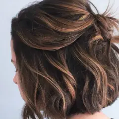 Coupe courte ondulée avec des reflets blonds subtils, le dessus est relevé en un chignon flou.
