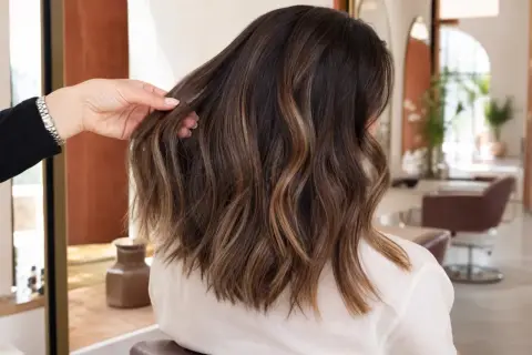 Une main ajuste une chevelure brune aux reflets subtils de balayage caramel, dans un salon de coiffure lumineux.