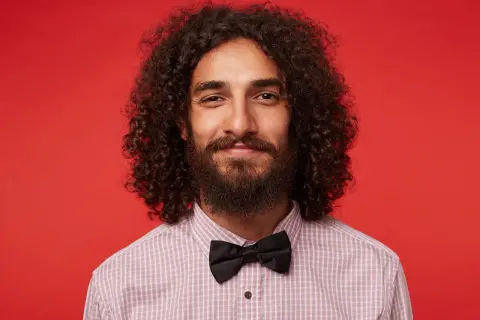 Homme souriant avec de longs cheveux bouclés, barbe et nœud papillon noir, sur fond rouge.