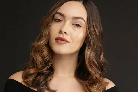 Une femme aux cheveux bruns ondulés, avec des reflets blonds, pose pour une photo. Elle a un maquillage naturel et un sourire subtil. Elle semble avoir réussi à faire un balayage maison.