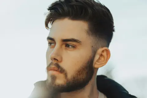Jeune homme avec une barbe courte et une coupe de cheveux dégradée, style taper fade. Il regarde au loin, le visage éclairé par une lumière douce.