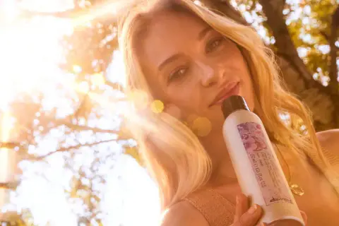 Une femme aux cheveux blonds, avec un balayage naturel, tient un produit Davines sous un soleil éclatant.