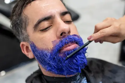 Un homme se fait teindre la barbe en bleu vif par une main tenant une brosse.