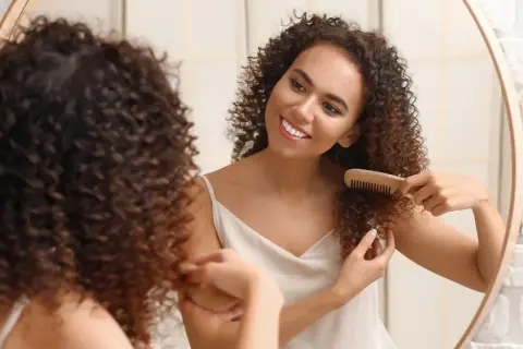 Une jeune femme souriante se regarde dans un miroir, peignant ses cheveux bouclés courts avec un peigne en bois.
