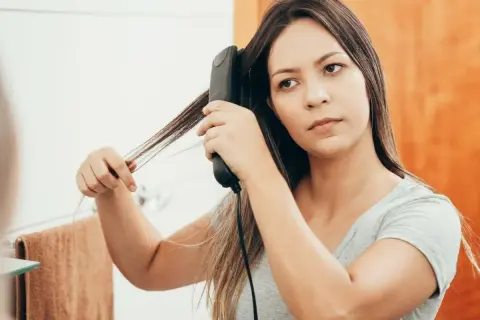 Une jeune femme réalise un lissage brésilien maison devant son miroir, utilisant un fer à lisser pour dompter ses cheveux.