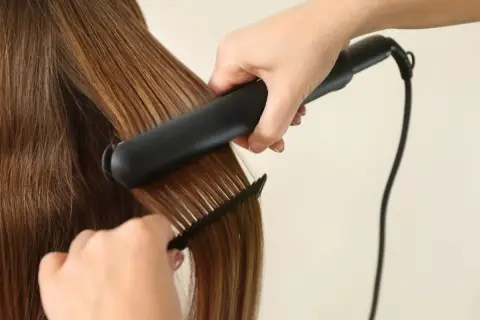 Une main utilise un fer à lisser pour lisser des cheveux bruns brillants, tandis qu'une autre main tient un peigne pour guider la mèche.