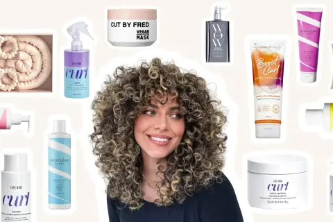 Une femme aux cheveux bouclés souriante, entourée de produits pour une routine cheveux bouclés : sprays, masques, gels et conditionneurs.