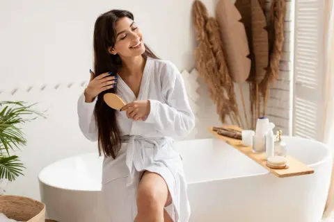 Une femme souriante en peignoir blanc se brosse les cheveux longs et bruns, un geste doux pour nourrir ses cheveux.