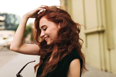 Une jeune femme aux cheveux roux, souriante, joue avec sa chevelure. Le **dosage coloration oxydant 30** a révélé cette belle teinte.