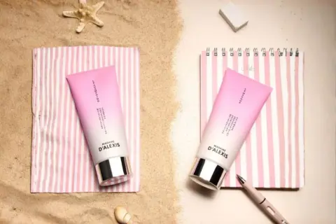 Deux tubes de crème rose sur des serviettes rayées, parfaits pour hydrater les cheveux sec après une journée à la plage.