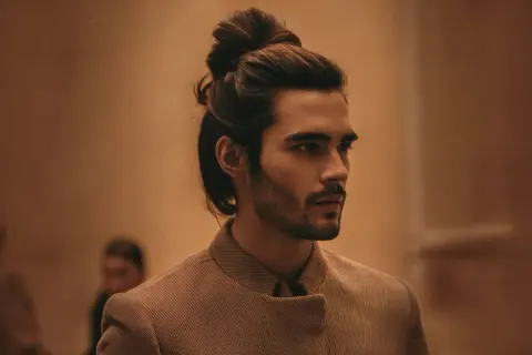 Un homme au chignon homme stylé, portant une veste texturée, regarde au loin.