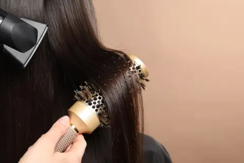 Une main utilise une brosse ronde pour coiffer des cheveux bruns, tandis qu'un sèche-cheveux souffle de l'air. Le brushing boucle est en cours.