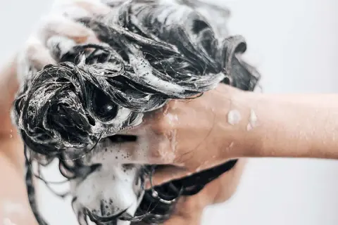 Une main savonne des cheveux sombres et mouillés, créant une mousse abondante. Une question se pose : peut-on se laver les cheveux au savon de Marseille ?