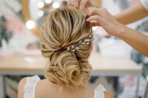 Une mariée se fait coiffer un chignon bas flou, orné d'une barrette scintillante. Les mains d'une coiffeuse ajustent les mèches.