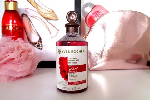 Bouteille de vinaigre de framboise pour cheveux Yves Rocher, avec des chaussures stylisées et une trousse de toilette rose en arrière-plan.