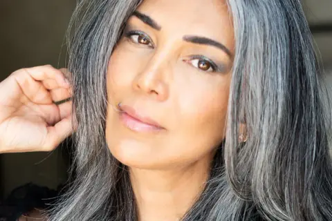 Une femme avec des cheveux gris mi-courts, le regard intense, touche doucement sa chevelure.