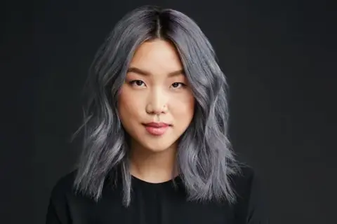 Une jeune femme au teint clair arbore une chevelure mi-longue, ondulée et d'un magnifique gris argenté. Elle porte un haut noir.