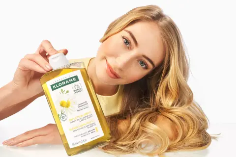 Une femme aux cheveux blonds tient un flacon de shampoing Klorane à la camomille, un produit pour cheveux éclaircissant.