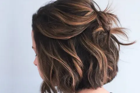 Coupe courte ondulée avec des reflets blonds subtils, le dessus est relevé en un chignon flou.