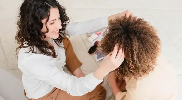 Deux femmes souriantes appliquent un bain d'huile sur des cheveux bouclés, un moment de soin capillaire, que les cheveux soient propres ou sales.