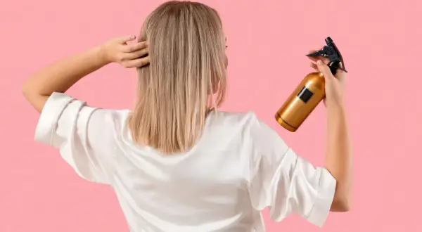 Une femme en peignoir blanc tient un flacon doré, prêt à utiliser du peroxyde d'hydrogène pour éclaircir ses cheveux blonds.